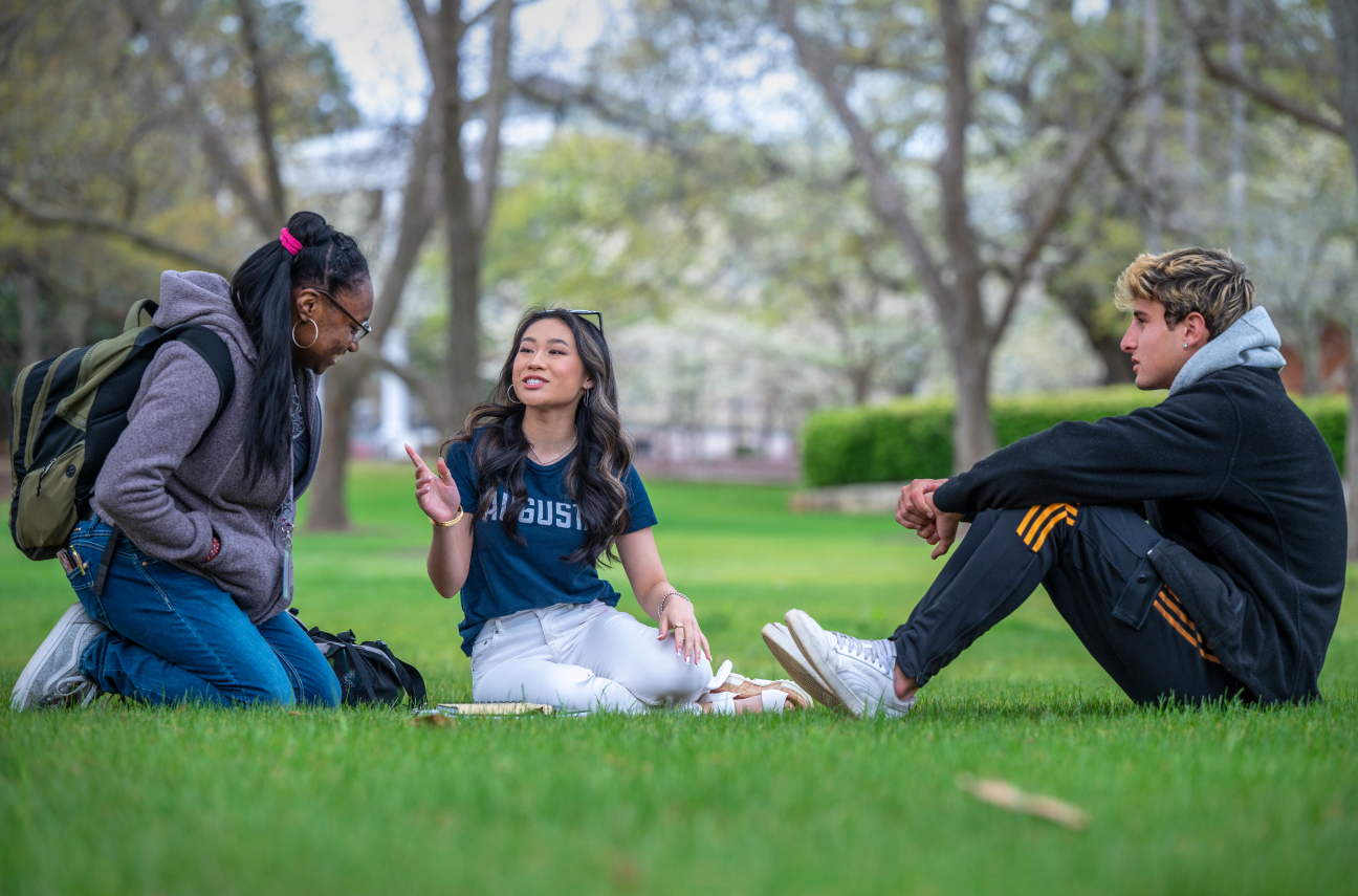 Three students sitting on the ground having a conversation on a green grassy lawn